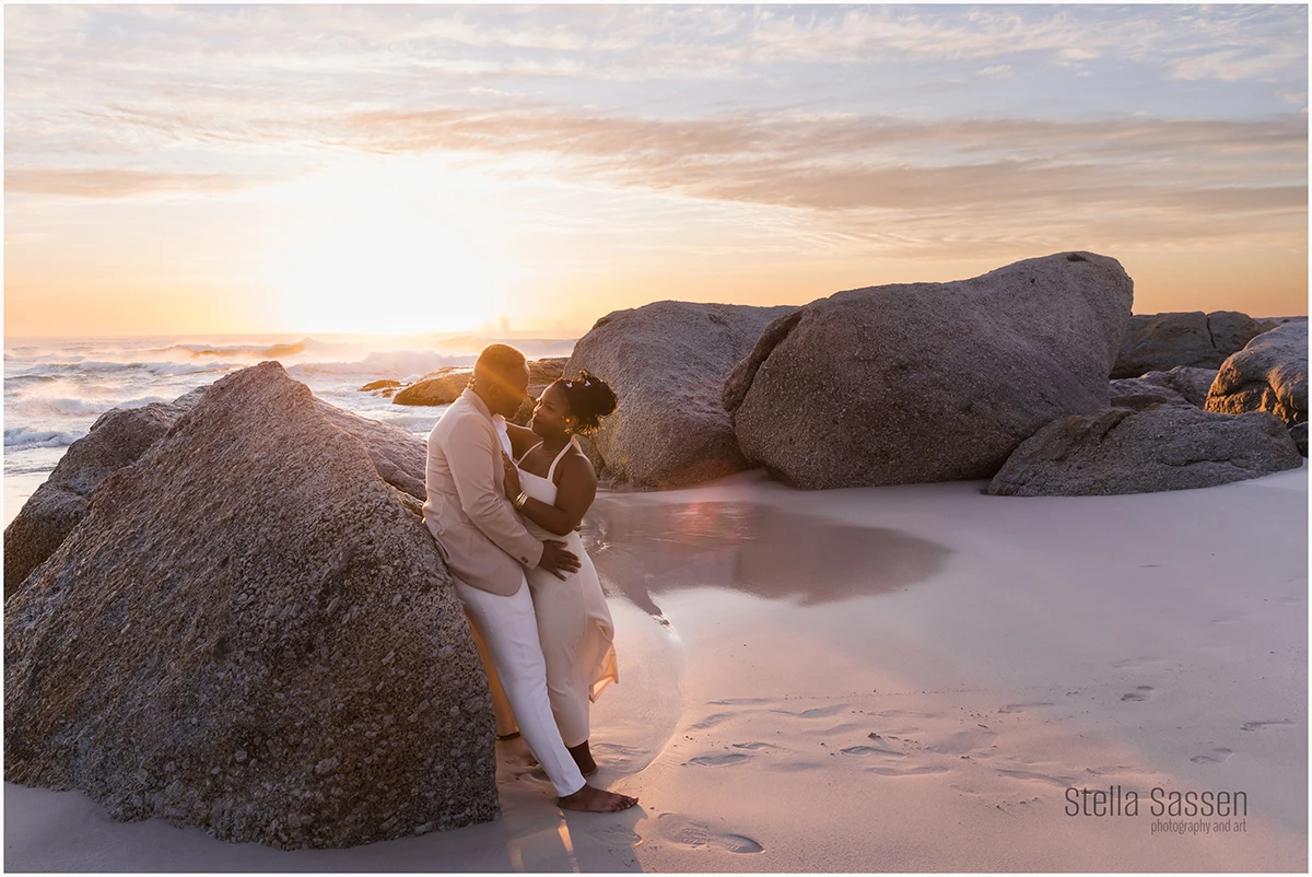 Couple embracing on a beach at sunset, standing among large granite rocks with soft waves and golden light in the background. The woman wears a white dress and the man a light suit, both barefoot on the wet sand. A romantic and intimate moment captured in the warm evening glow