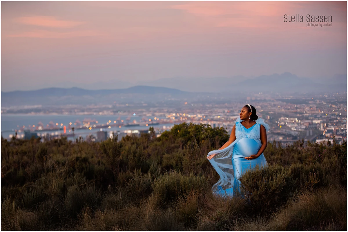An expectant mother stands on Signal Hill at sunset, wearing a light blue dress. She holds her belly and looks to the side, with Cape Town city lights and distant mountains in the background. The scene is calm and natural, capturing a quiet moment as day turns to night.