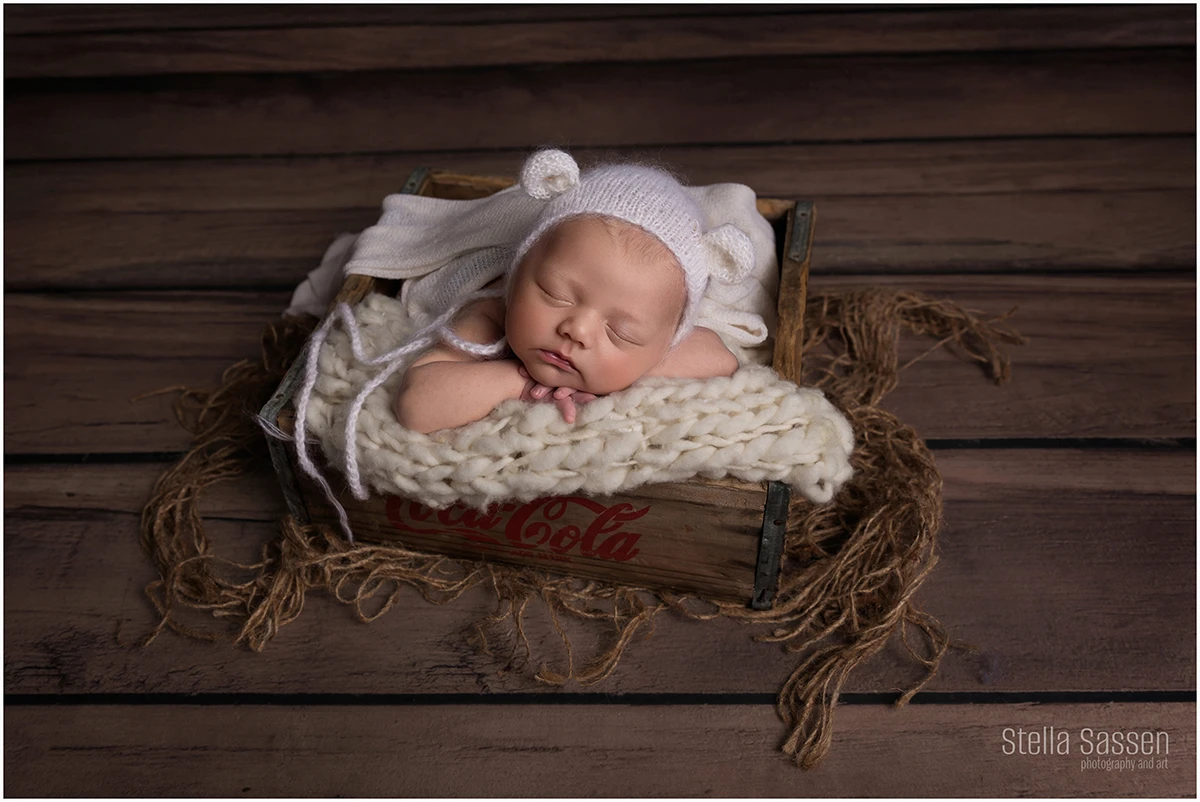 A peaceful newborn moment captured in rich, earthy tones. This little one is nestled in a vintage Coca-Cola crate, resting on a chunky cream knit blanket. Wearing a soft white bonnet with bear ears, baby sleeps soundly with hands tucked under their chin. 