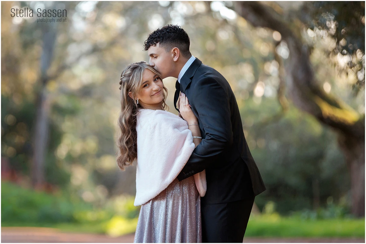 A young couple dressed for a matric ball stands outdoors, surrounded by soft, natural light and blurred greenery. The young woman, in a shimmering silver dress and a light pink shawl, smiles gently at the camera while the young man, in a classic black suit, leans down to kiss her forehead. The moment feels warm, elegant, and affectionate.