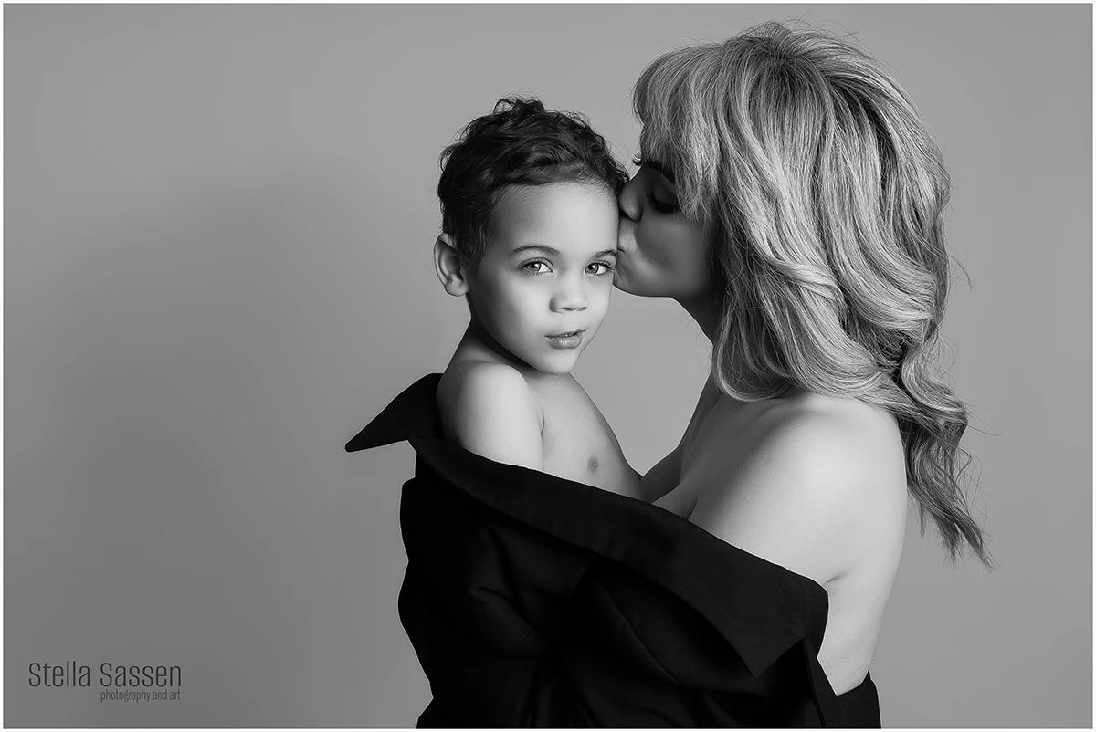 A black and white studio portrait of a mother holding her young son close. The mother kisses him gently on the temple while he looks directly into the camera. Wrapped in a draped black fabric, the image highlights intimacy, love, and the timeless bond between mother and child.