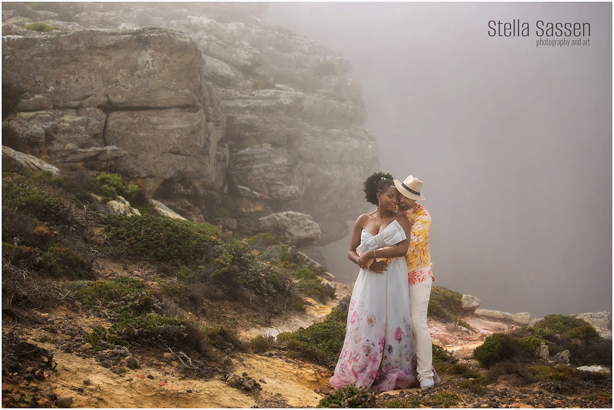 A couple stands on a rocky cliff surrounded by mist and fog, creating a dreamy and atmospheric scene. The pair is dressed elegantly, with the woman’s flowing dress adding movement against the rugged, textured rocks. The vast foggy backdrop gives a sense of mystery and isolation