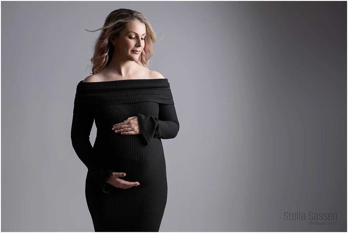 A studio maternity portrait of a confident pregnant woman in a tight fitting black dress, highlighting the belly
